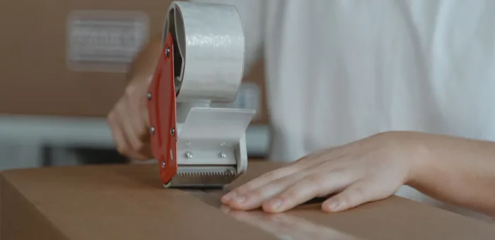 A hand running a tape gun along the seam of a carton at the packing bench