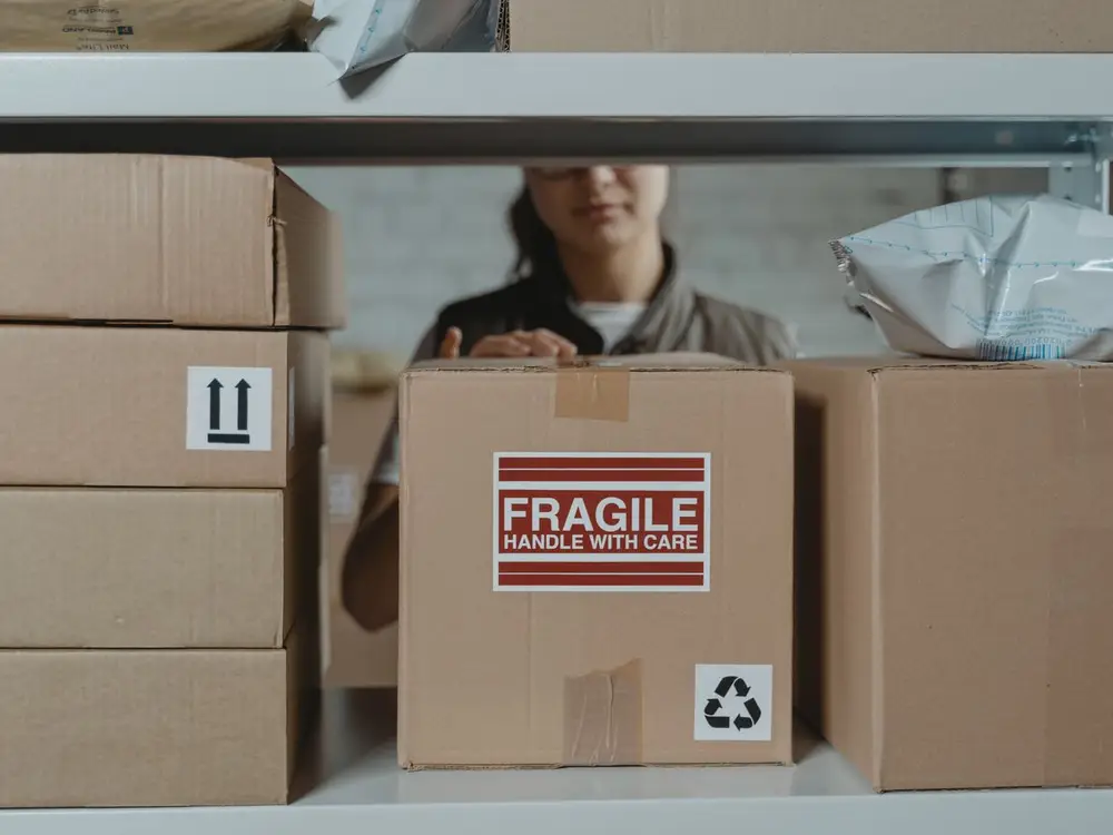 Cartons with fragile and this-way-up handling labels on warehouse shelving, a worker checking a box behind